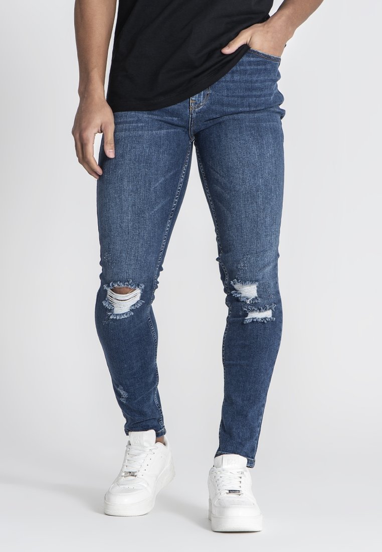Jeans ajustados de denim azul oscuro con desgaste en las rodillas, combinados con zapatillas blancas. Cuenta con bolsillos estándar y pasadores para cinturón.