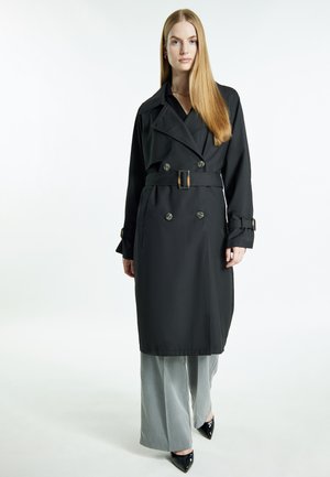 Donna con capelli lunghi biondi che indossa un trench nero con cintura, pantaloni grigi a gamba larga e scarpe nere appuntite, in piedi davanti a uno sfondo bianco.