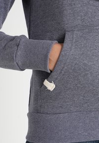 Szara bluza z prążkowanymi mankietami i ściągaczem u dołu, z boczną kieszenią ozdobioną kremową metką kontrastującą z resztą. Materiał wygląda na miękki i o fakturze.