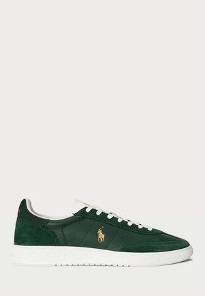 Groene suède en leren sneakers met een witte rubberen zool, voorzien van een gouden logo aan de zijkant en witte veters. Klassiek laag ontwerp.