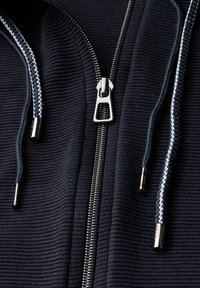 Schwarze Zip-Hoodie aus strukturiertem Gewebe. Verfügt über einen silbernen Reißverschluss, kontrastierende weiße und schwarze Kordeln sowie elegante metallische Hardware-Details.