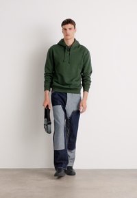 Felpa verde, jeans in denim con toppe a contrasto in grigio e blu scuro, scarpe nere e una borsa a tracolla grigia. Sfondo semplice.