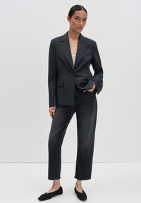 Vrouw in een zwart blazer, witte top, vervaagde zwarte jeans en zwarte platte schoenen staat tegen een effen lichtgrijze achtergrond.