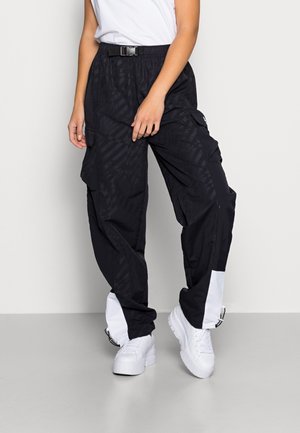 Osoba ubrana w czarne spodnie typu cargo z bocznymi kieszeniami i białymi panelami przy kostkach, do tego białe sneakersy na platformie oraz biały top.