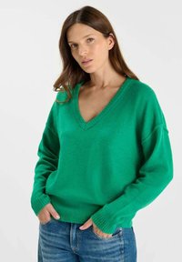 Groene, gebreide trui met een v-hals, verlaagde schouders en geribde manchetten, gecombineerd met blauwe spijkerbroeken en een gladde textuur.