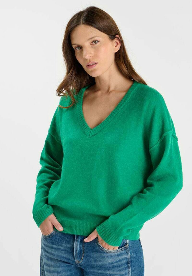 Groene, gebreide trui met een v-hals, verlaagde schouders en geribde manchetten, gecombineerd met blauwe spijkerbroeken en een gladde textuur.