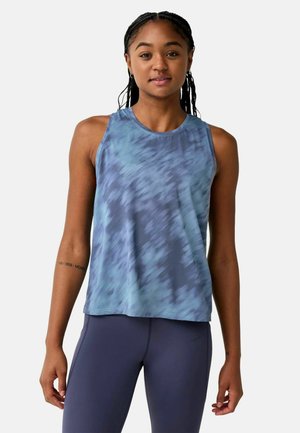Jonge vrouw met ingevlochten haar, gekleed in een blauw tie-dye mouwloos topje en donkerblauwe legging, staand tegen een effen achtergrond.