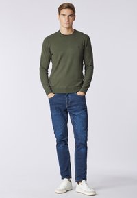 Pullover in maglia verde oliva con scollo rotondo, abbinato a jeans blu in denim e sneakers bianche. Il pullover presenta un piccolo logo sul petto.