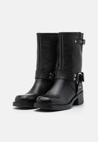 Botas de tobillo de cuero negro con un acabado suave, una correa con herrajes plateados y un tacón bajo apilado. Detalles de costura visibles.