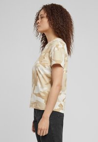T-shirt a maniche corte, beige con motivo camouflage, in misto cotone. Presenta un collo rotondo, vestibilità comoda e una texture leggera. Abbinato a pantaloni grigio scuro.