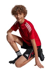 Rode atletische jersey met witte strepen, zwarte shorts met rode en witte accenten, zwarte voetbalschoenen en gestreepte zwart-witte sokken.