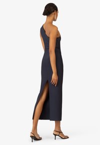 Robe maxi ajustée bleu marine à épaule unique, avec une fente sur le côté et une texture lisse. Portée avec des talons noirs à lanières.