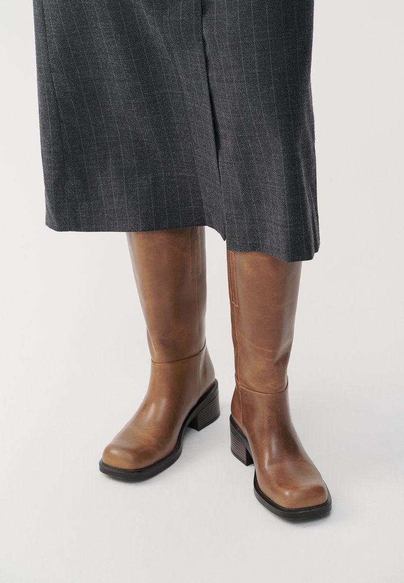 Stivali alti fino al ginocchio in pelle marrone con punta quadrata, tacco spesso nero e superficie liscia, abbinati a pantaloni grigi a righe.