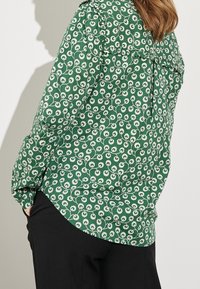 Blouse florale verte avec des motifs de fleurs blanches, tissu doux, manches longues, détail des épaules froncé, ourlet arrondi, associée à un pantalon noir.