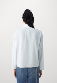 Blusa in satin azzurro chiaro con maniche lunghe, vestibilità morbida e spacco posteriore. Presenta una lucentezza sottile e un orlo arrotondato.