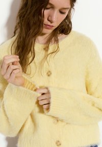 Gelber, samtweicher Cardigan aus weichem Material, mit Rundhalsausschnitt, langen Ärmeln und fünf Knöpfen auf der Vorderseite.