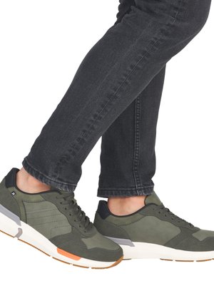 Green suede sneakers with black accents, lace-up design, cushioned white sole, and orange detail on the toe. Worn with dark jeans.