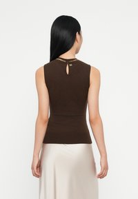 Bruine mouwloze top met een getailleerd ontwerp, geribbelde textuur en een gouden kettingaccent bij de halslijn. Details met een sleutelgat aan de achterkant.
