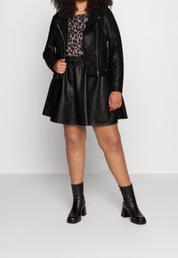 Svart läderbikerjacka över en leopardmönstrad topp, kombinerad med en svart fusk läderkjol och svarta klackade ankelstövlar, framifrån.