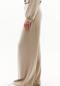 Pantalones de pierna ancha en beige, hechos de una tela suave, con una textura lisa y una costura lateral delgada. El diseño incluye una cintura alta y un ajuste relajado.