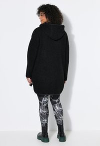 Mujer con cabello rizado que lleva un abrigo negro con capucha, leggings florales en blanco y negro y botas negras con suelas verdes, de pie mirando hacia atrás.