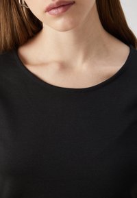 Czarna bawełniana koszulka z okrągłym dekoltem, gładką teksturą i krótkimi rękawami, obnażająca górną część klatki piersiowej i linię żuchwy.