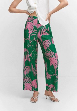 Frau trägt hoch taillierte, weit geschnittene Hosen mit grünem und pinkem tropischem Blättermuster, kombiniert mit weißem, ärmellosem, geknöpftem Oberteil und Sandalen mit Absatz.