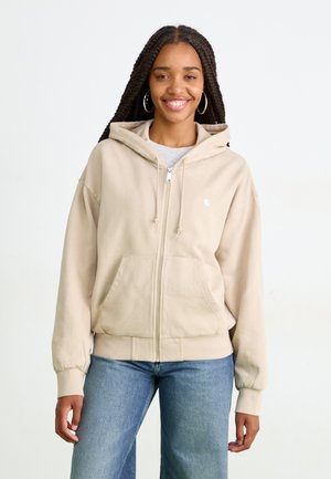 Beigā krāsā zip-up kapuce ar priekšējo kabatu un ribotām piedurknēm. Iespējo kapuci un mazu logotipu uz krūtīm. Valkāta ar gaismas zilām platām biksēm.