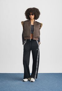 Femme portant une veste sans manches marron foncé avec bordure en fourrure, un crop top noir, un pantalon de survêtement noir avec des bandes blanches, des baskets blanches et des lunettes de soleil.