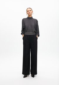 Black and white polka dot blouse with a high collar, long sleeves, and an elastic band at the waist; paired with wide-leg black trousers.