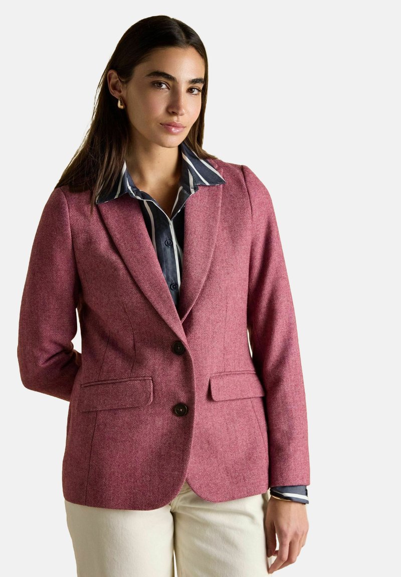 Žena s rovnými hnedými vlasmi má na sebe zlaté náušnice, bordový blejzer cez pruhovanú námornícku blúzku a krémové nohavice, pózuje na bielom pozadí.