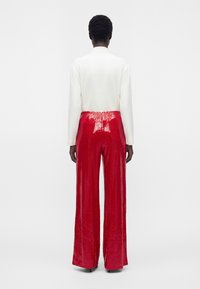 Personne de dos, portant un haut blanc à manches longues et un pantalon rouge brillant à jambes larges, sur un fond clair et uni.