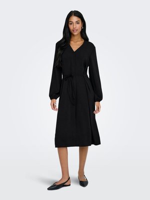Femme debout portant une robe midi noire à manches longues, col en V et ceinture taille, associée à des ballerines noires pointues à bride arrière.
