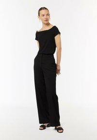 Czarna bluzka bez ramiączek, szerokie czarne spodnie i czarne sandały z plecionym wzorem. Prosta, elegancka sylwetka z gładką teksturą materiału.