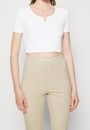 Kobieta w białej, krótkiej bluzce z krótkim rękawem i beżowych, wysokich spodniach z logo 'Calvin Klein Jeans' na pasku.