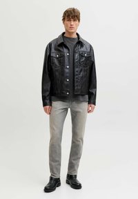 Chaqueta de cuero sintético negro con botones de presión y dos bolsillos en el pecho, combinada con jeans de mezclilla gris y botines negros. La modelo está de pie contra un fondo liso.