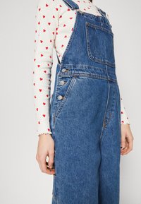 Salopette in denim con gambe larghe, di colore blu, spalline regolabili e dettagli in bottoni argentati. Indossata sopra una camicia bianca a maniche lunghe con cuori rossi.