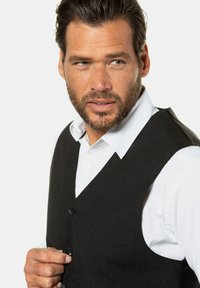 Chaleco negro con diseño de escote en V, que presenta cuatro botones y una tela texturizada. Lleva puesto sobre una camisa blanca con cuello para crear contraste.