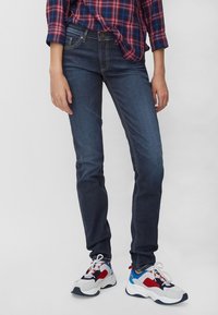 Pantalones vaqueros skinny de denim azul oscuro con sutiles desgastes, bolsillos frontales y una cintura de altura media, combinados con zapatillas blancas, rojas y grises.