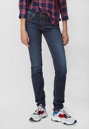 Pantalones vaqueros skinny de denim azul oscuro con sutiles desgastes, bolsillos frontales y una cintura de altura media, combinados con zapatillas blancas, rojas y grises.