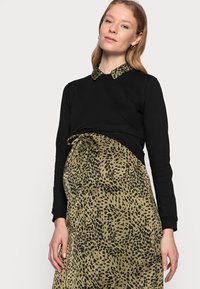 Top preto de mangas longas com um vestido verde estampado de leopardo por baixo, apresentando uma cintura amarrada e detalhes em botão. Textura suave e design em camadas.