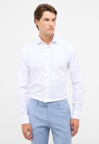 Mann in einem enganliegenden weißen Hemd und hellblauen Hosen, stehend mit entspannten Händen an den Seiten, schaut nach rechts.
