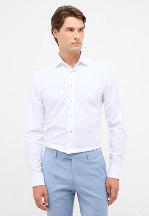 Mann in einem enganliegenden weißen Hemd und hellblauen Hosen, stehend mit entspannten Händen an den Seiten, schaut nach rechts.