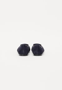 Mocassins en suede bleu marine avec une texture lisse, présentant un bout arrondi et un accent de nœud décoratif sur la partie supérieure. Semelles en caoutchouc noir.