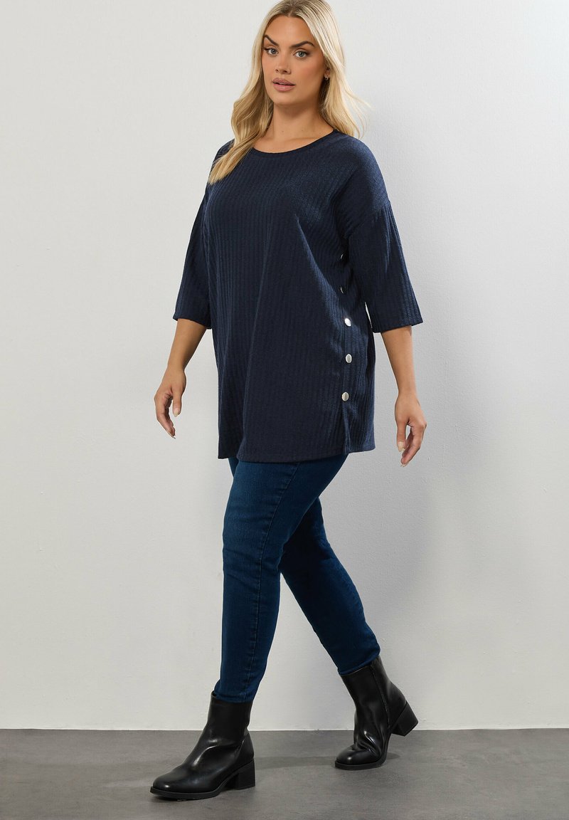 Top a coste blu navy con maniche a tre quarti, bottoni laterali e una vestibilità comoda, abbinato a jeans skinny blu e stivali neri alla caviglia.