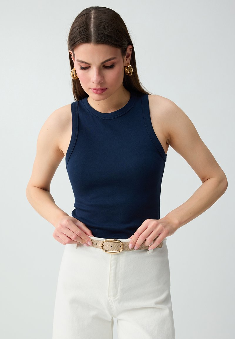 Femme aux cheveux bruns et raides portant des boucles d'oreilles créoles dorées, un haut sans manches bleu marine, un pantalon blanc, ajustant une ceinture beige avec une boucle dorée.