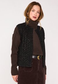 Gilet nero a maglia con motivi texture, indossato sopra un maglione marrone a collo alto. L'outfit è completato da una gonna in velluto a coste marrone e una cintura nera.