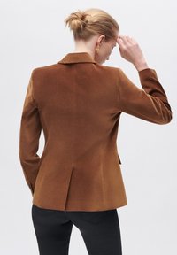 Brauner maßgeschneiderter Blazer mit Reverskragen, einem Schlitz hinten und langen Ärmeln, aus einem glatten Stoff gefertigt. Kombination mit schwarzen Hosen.