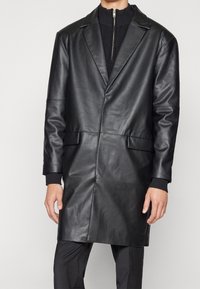 Czarny długi płaszcz skórzany z klapami z wycięciem, dwoma przednimi kieszeniami i gładką fakturą. Noszony na czarnym swetrze z wysokim kołnierzem.