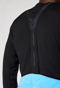 Juodas ilgomis rankovėmis marškinėliai su reguliuojamomis juodomis petnešomis, ant kurių yra logotipas, dėvimi su dangaus mėlynumo prijuoste su lygiu paviršiumi ir siūlėmis.